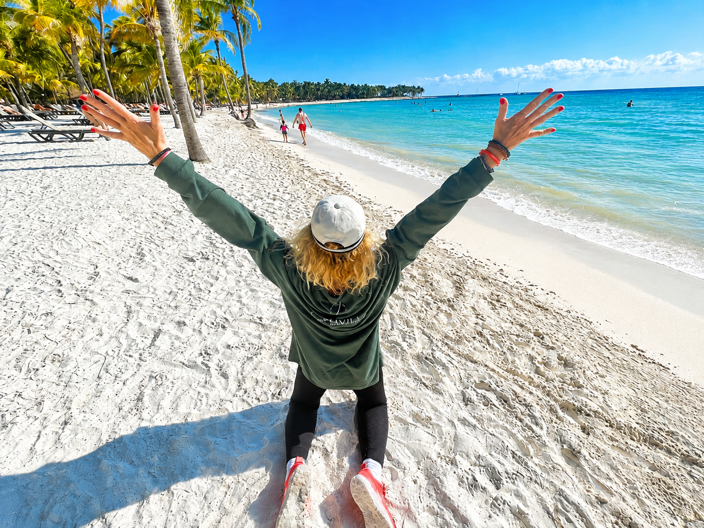Dos tourné face à la mer turquoise à Cancún, Liliane Clavel Pardo lève les bras après une walking meditation durant une retraite de Joe Dispenza au Barceló Maya en 2022. Cheveux bouclés coupés au carré, posture d’ouverture corporelle, plage tropicale et sensation d’expansion physique